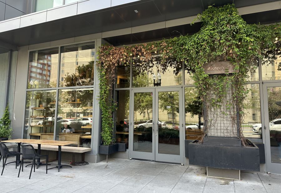 The image shows the exterior of a café named *A Baked Joint*, featuring modern glass windows, greenery along the entrance, and outdoor seating on the sidewalk.
